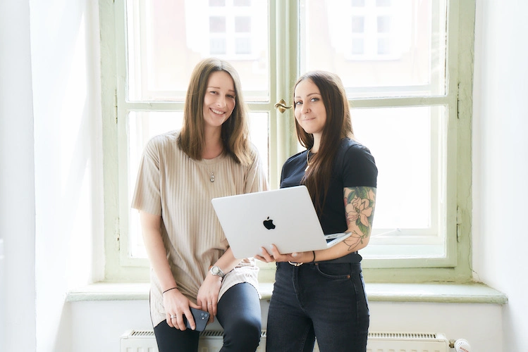 Ina und Tamara von Frida Grün stehen im Büro vor einem Fenster,lächeln und halten einen Laptop in der Hand