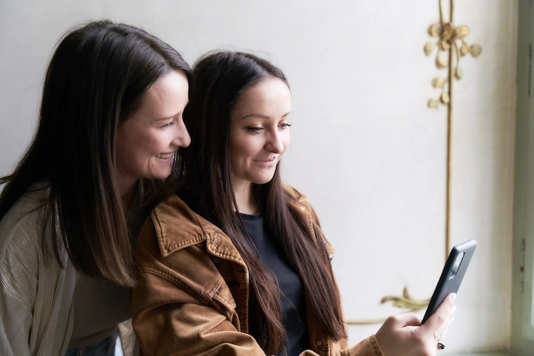 Tamara Gosch und Sabrina Egger-Feiel von FRIDA GRÜN stehen nebeneinander und blicken gemeinsam auf ein Handy. Sie tragen Jacken und lächeln.