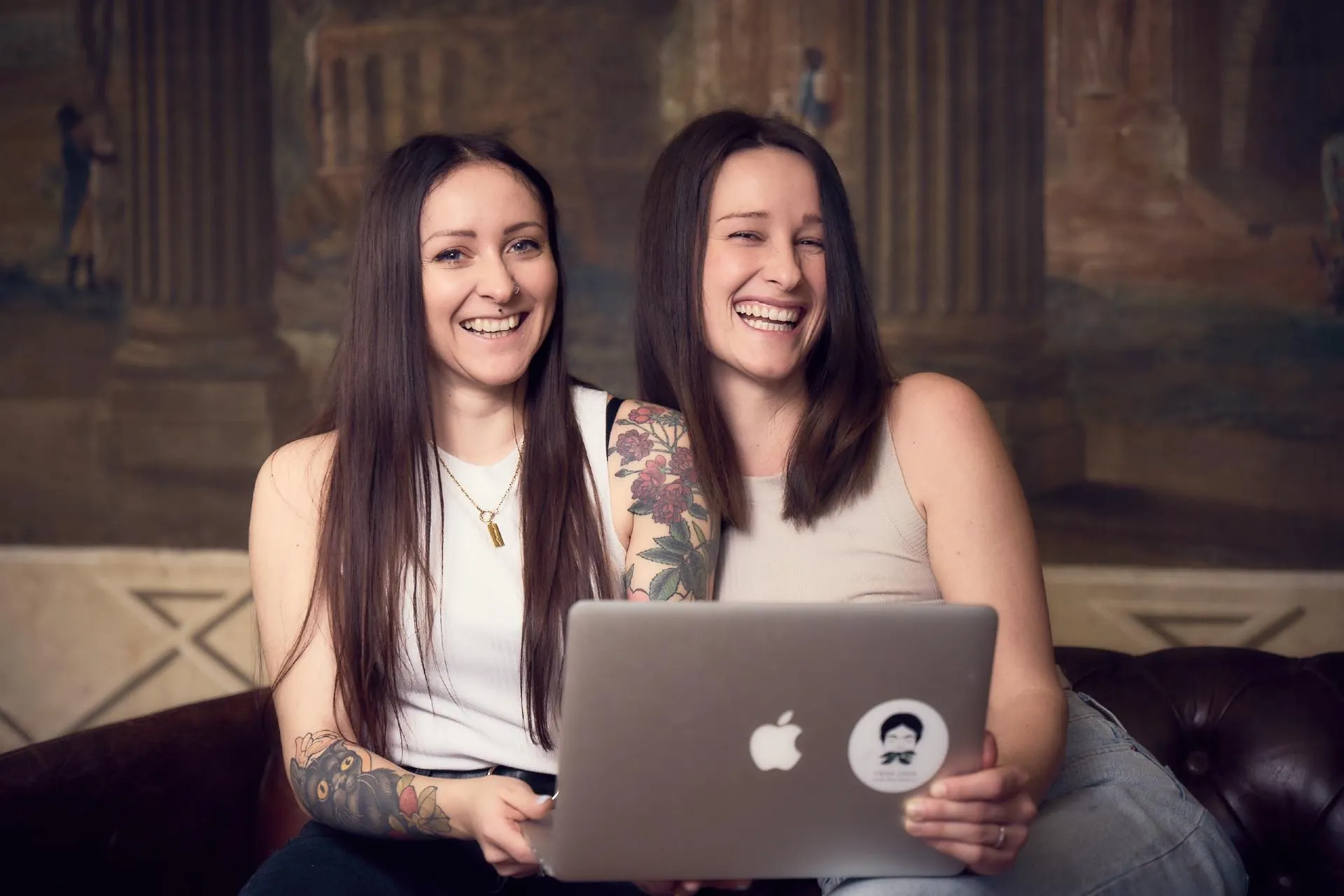 Ina und Tamara von FRIDA GRÜN sitzen mit einem Laptop auf einer Couch und lächeln in die Kamera